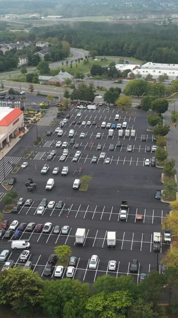 Aerial view of a large shopping-centre parking lot filled with cars, bordered by roads and green trees.