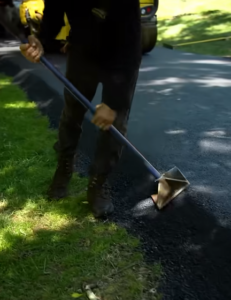 Worker in dark clothing and boots using a hand tamper to compact freshly laid asphalt beside a driveway.