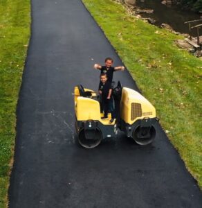 Two children ride a yellow road roller on a park path beside a grassy verge and a stream