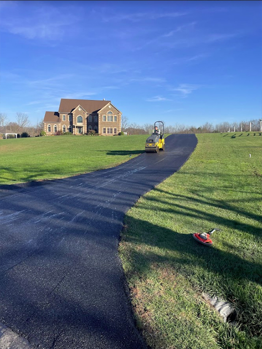Curved freshly paved driveway leading to a large brick house; a yellow paving roller is on the road.