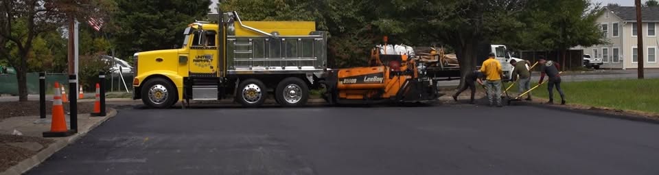 Road construction scene with a yellow dump truck and orange asphalt paver laying new pavement while workers guide the process.